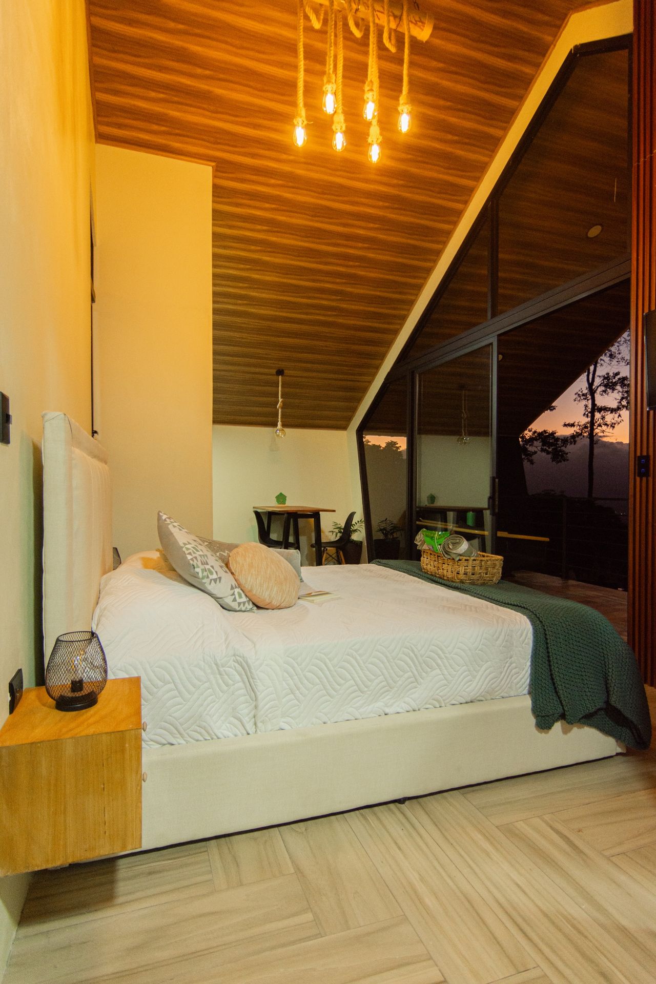 Habitación de noche con cama Queen, lámparas cálidas y ventanal al atardecer